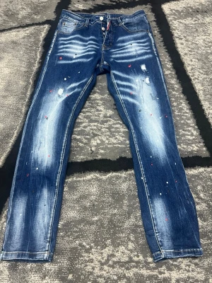 Dsquared2 blå slim jeans med patch - Säljer ett par blå slim jeans från Dsquared2 med coola slitningar, färgstänk och en stor gul-röd patch med nummer 2 på bakfickan. Jeansen har klassisk femficksdesign och är tillverkade i bomull. Perfekt för dig som vill sticka ut med din stil.