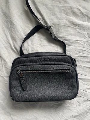 Svart Michael Kors väska - Snygg svart axelväska från Michael Kors med diskret mönster och silverfärgade dragkedjor. Väskan har flera fack, justerbar rem och är tillverkad i skinn med präglad yta. Perfekt storlek för att bära med sig det viktigaste och har en modern look.