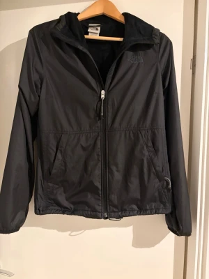 Svart vindjacka från The North Face - Säljer en svart vindjacka från The North Face med huva och dragkedja framtill. Jackan har två fickor med dragkedja och mjukt fleecefoder på insidan. Perfekt för blåsiga dagar och enkel att matcha med allt. Använd bara ett par gånger