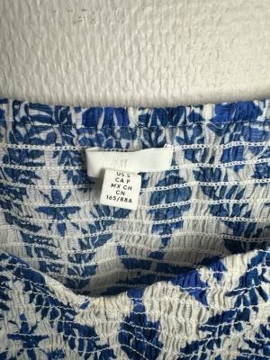 Blommig blus med puffärm från H&M - Säljer en vit och blå blommig blus från H&M i storlek S. Blusen har puffiga korta ärmar, smockad kropp och fyrkantig halsringning. Materialet är lätt och luftigt, perfekt för varmare dagar. Mönstret är i djupblå toner med blommiga detaljer.