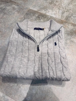 Grå stickad half zip tröja Polo Ralph Lauren - Säljer en grå kabelstickad tröja från Polo Ralph Lauren i storlek S. Tröjan har half zip med läderdetalj, ribbad krage och muddar samt broderad logga på bröstet. Perfekt för dig som gillar klassisk och stilren design. Den är helt ny och kostar 3000 kr i nypris, säljer för att den är lite för liten på mig.