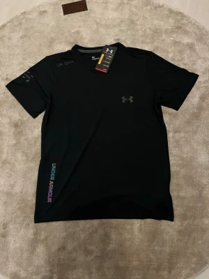 Svart Under Armour tränings-tshirt - Säljer en svart Under Armour Threadborne HeatGear t-shirt med reflekterande logga och detaljer. Materialet är lätt och syntetiskt, perfekt för träning. UA-Spec och Under Armour-tryck på ärm och nederkant. Snygg passform och modern look.