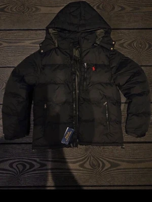 Svart pufferjacka från Polo Ralph Lauren - Svart pufferjacka från Polo Ralph Lauren med huva och dragkedjor på fickorna. Jackan har ett rött Polo-logo broderat på bröstet och quiltad design. Perfekt för kalla dagar och har en snygg, sportig vibe.  Priset kan diskuteras vid snabb affär!  