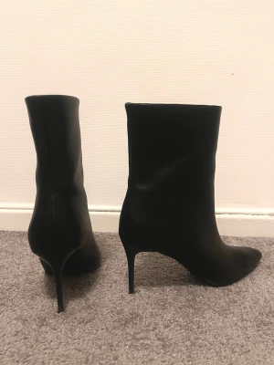 Svarta stövletter med spetsig tå och klack - Säljer ett par svarta stövletter i skinn med elegant spetsig tå och hög smal klack. Snyggt minimalistisk design med diskret YSL-logga i guld på hälen. Perfekta för dig som gillar stilrena boots med lyxig känsla.