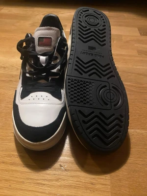 Svarta och vita sneakers från Polo Sport - Snygga sneakers från Polo Sport i svart och vitt med klassisk låg profil. Skorna har svart snörning, rund tå och en cool amerikansk flagga på plösen. Materialet är en mix av skinn och syntet, med kontrasterande detaljer och Polo Sport-logga på sidan.