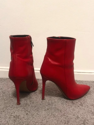 Röda stövletter med spetsig tå - Säljer ett par röda stövletter från Ognibene, tillverkade i Italien. Skorna har spetsig tå, hög klack och dragkedja på sidan. Materialet är slätt skinn och färgen är intensiv röd. Perfekt för dig som vill sticka ut med en cool look.