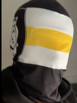AIK mask - Säljer en unik balaclava i svart, vitt och gult med ett tryckt ansikte på sidan. Masken har en gul mesh-detalj över ögonen och är tillverkad i ett syntetmaterial som känns mjukt och följsamt. Perfekt för dig som vill sticka ut med din stil. AIK MASK