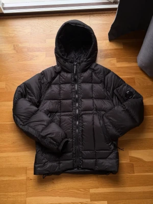 Svart pufferjacka från C.P. Company - Säljer en svart pufferjacka från C.P. Company med huva och ikonisk linsdetalj på ärmen. Jackan har quiltad design, dragkedja framtill och är tillverkad i lätt, syntetiskt material. Perfektum för kalla dagar och riktigt snygg streetwear-vibe.