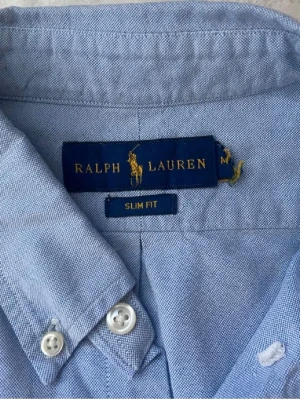 Blå slim fit skjorta från Ralph Lauren - Snygg ljusblå skjorta från Ralph Lauren i slim fit-modell. Klassisk button-down krage och broderad logga på bröstet. Skjortan är tillverkad i mjuk bomull och har vita knappar framtill. Perfekt för dig som gillar stilrena och tidlösa plagg.