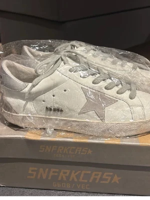 Vita sneakers från Golden Goose - Snygga vita sneakers från Golden Goose med den ikoniska stjärnan på sidan och grå snörning. Skorna har en vintage-inspirerad look med slitna detaljer och är tillverkade i skinn. Perfekta för dig som gillar en avslappnad men trendig stil.