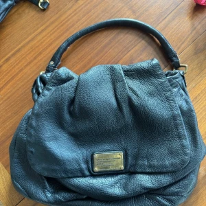 Svart skinnväska från Marc by Marc Jacobs - Säljer en snygg svart skinnväska från Marc by Marc Jacobs. Väskan har en stilren design med en metallplatta framtill och ett mönstrat foder. Insidan är tvättad men kommer inte bli vitare än på bild, men har tydliga spår av användning. Nött på metall och handtag men har mycket kvar att ge. Cirka 40 bred och 25 hög. 