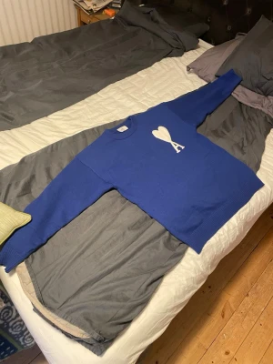 Blå stickad tröja från Ami Paris - Säljer en blå stickad tröja från Ami Paris med vitt hjärta och A på bröstet. Tröjan har rund hals, ribbade muddar och är långärmad. Materialet är 85% ekologisk bomull och 15% ull, vilket gör den både mjuk och skön. Perfekt för dig som gillar stilrena detaljer.