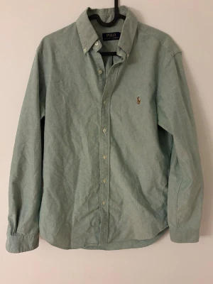Grön skjorta från Polo Ralph Lauren - Snygg grön skjorta från Polo Ralph Lauren med klassisk krage och knappar hela vägen. Broderad logga med polospelare på bröstet. Skjortan är långärmad och tillverkad i mjuk bomull, perfekt för en chill och stilren look.