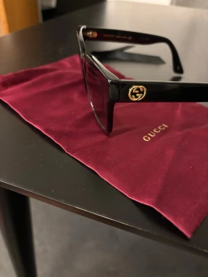 Svart Gucci solglasögon med guldlogo - Snygga svart solglasögon från Gucci med klassisk fyrkantig form och guldfärgad logodetalj på sidan. Perfekt för dig som vill ha en exklusiv och trendig look.