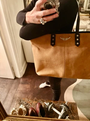 Beige mocka axelväska med svarta detaljer - Säljer en snygg beige axelväska från Zadig & Voltaire i mocka med svarta handtag och silverfärgade nitar. Väskan har ett stilrent emblem framtill och är perfekt för dig som gillar klassisk design med en cool touch.