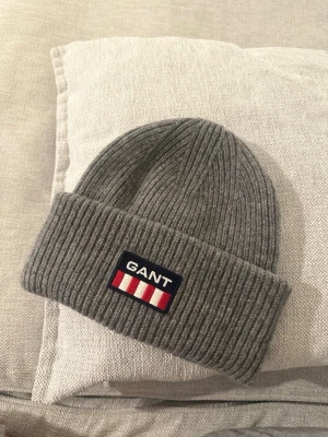 GANT grå ull mössa - Säljer en snygg grå ribbstickad mössa från GANT med uppvikt kant och klassisk logga framtill. Mössan är i mjukt material och passar perfekt för kyliga dagar. Enkel och stilren design som funkar till det mesta.