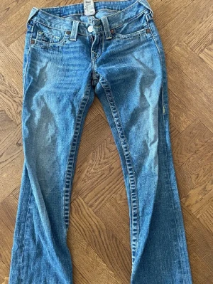 True Religion blå jeans bootcut - Säljer ett par klassiska blå jeans från True Religion med bootcut-snitt och kontrastsömmar. Jeansen har fem fickor, låg midja och är tillverkade i slitstarkt denim med gul söm. Märkespatch bak på linningen och coola detaljer på bakfickan.