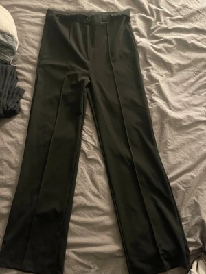 Svarta kostymbyxor med raka ben - Snygga svarta kostymbyxor med klassisk rak passform och pressveck framtill. Byxorna har hög midja och stängs med knapp och dragkedja. Perfekta för en stilren look och passar till många olika outfits. Storlek S - M