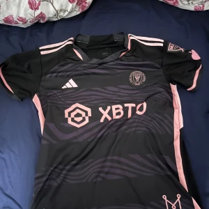 Inter Miami Messi 10 matchtröja - Säljer en svart och rosa Inter Miami matchtröja från Adidas med Messi och nummer 10 på ryggen. Tröjan har zebramönster, rosa detaljer och klubbmärke på bröstet. Perfekt för dig som älskar fotboll och vill ha en snygg supportertröja.