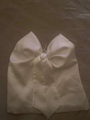 Vit bandeau topp med stor rosett - Supersnygg vit bandeau topp med en stor rosett framtill som verkligen drar blickarna till sig. Toppens material är satin och känns lyxigt, perfekt för att sticka ut. Axelbandslös design som ger en clean och modern look. Helt ny, endast testad!