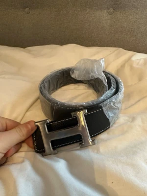 Svart skärp med H-spänne i metall - Snyggt svart skärp med grov textur och ett stort H-format spänne i silverfärgad metall. Skärpet har vita sömmar längs kanterna och är tillverkat i skinn. Perfekt för att ge outfiten en extra touch.