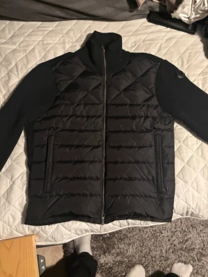 Svart Moncler pufferjacka med stickade ärmar - Säljer en svart pufferjacka från Moncler med stickade ärmar och hög krage. Jackan har quiltad front, två fickor med dragkedja och snyggt Moncler-märke på ärmen. Materialmix av polyester och stickad ull ger en cool look. Perfekt för dig som vill ha något stilrent och bekvämt. ‼️ pris kan diskuteras‼️