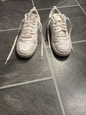 Nike sneakers med mintgrön swoosh - Fräscha vita Nike sneakers med mintgrön swoosh på sidorna och Nike-logga på hälen. Lågt skaft, rund tå och klassisk snörning. Skorna är tillverkade i skinn och har perforeringar på tån för extra ventilation. Perfekta för dig som gillar stilrena och sportiga sneakers. Fläckarna går bort med diskmedel och ljummet vatten