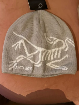 Grå mössa Arc'teryx med logga - Säljer en grå mössa från Arc'teryx med den ikoniska dinosaurieloggan i svart och vitt. Mössan är stickad och har en clean, enkel design som passar perfekt till kalla dagar. Materialet är mjukt och bekvämt.