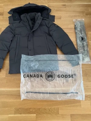 Svart pufferjacka från Canada Goose - Säljer en svart pufferjacka från Canada Goose med avtagbar huva och klassisk logga på ärmen. Jackan har långärmade ärmar, dragkedja och knappar framtill. Perfekt för kalla vinterdagar och har en clean, modern look.
