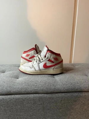 Nike Air Jordan 1 Mid vit/röd EUR 38 - Nike Air Jordan 1 Mid sneakers i vitt och rött med klassisk röd swoosh och detaljer. Skorna har snörning, rund tå och platt sula. Ovandelen är i skinn och mesh, med röd yttersula och Air Jordan-logga på sidan och plös. Perfekt för dig som gillar retrostil och streetwear.