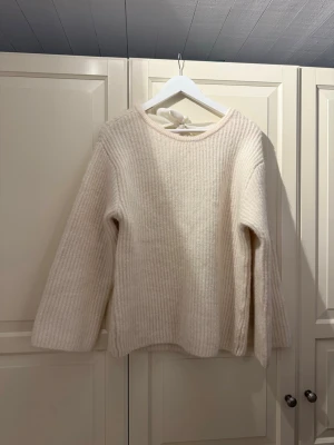 Beige stickad tröja med rosett - Superfin beige stickad tröja med långärmad modell och ribbad struktur. Tröjan har en öppen rygg med en stor rosett i nacken som ger en extra snygg detalj. Perfekt för dig som gillar mjuka och mysiga plagg med lite twist.