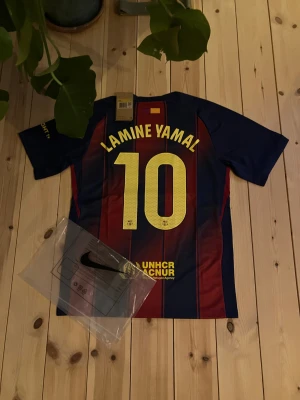 FC Barcelona tröja Lamine Yamal #10 - Säljer en officiell FC Barcelona matchtröja från Nike med Lamine Yamal och nummer 10 på ryggen. Tröjan har klassiska blå och röda ränder, klubbmärke och Spotify-logga på bröstet samt UNHCR-tryck. Perfekt för dig som älskar fotboll och Barca! Inte riktig men gättebra kvalitet