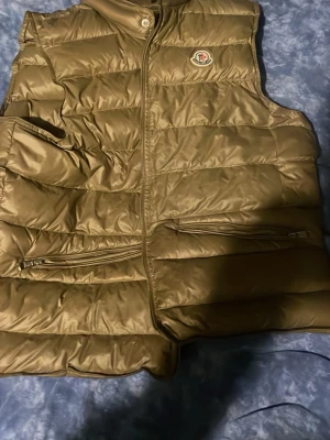Beige dunjacka från Moncler - Säljer en beige dunjacka från Moncler med quiltad design. Jackan har en glansig yta och är fylld med dun för extra värme. Perfekt för kalla dagar och har en stilren look som passar till många outfits.
