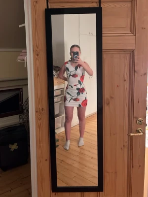 Vit kort klänning med röda rosor - Säljer en vit, tight kortklänning med stora röda rosor och gröna blad. Klänningen är ärmlös och har en figurnära passform som framhäver siluetten. Perfekt för dig som gillar färgstarka mönster och vill sticka ut. Fråga mig om ni undrar något 😊