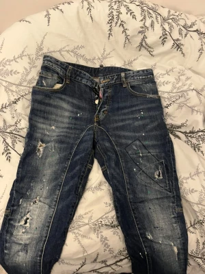 Dsquared2 Classic Kenny jeans med dragkedja - Säljer ett par Dsquared2 Classic Kenny Twist jeans i mörkblå tvätt med slitningar, färgstänk och coola detaljer. Byxorna har en synlig dragkedja framtill, flera fickor och grova sömmar. Märke på bakfickan och tillverkade i Italien. Riktigt street och edgy vibe.