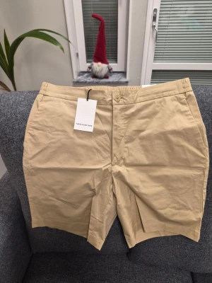THESE GLORY DAYS T Adel Poplin beigea shorts, storlek 48 - Snygga beige shorts från These Glory Days med elastisk midja, snörning och dragkedja framtill. Modellen har raka ben och två bakfickor. Materialet är mjuk bomull som känns skönt mot huden. Perfekta för varma dagar och enkel att matcha med t-shirt eller skjorta.