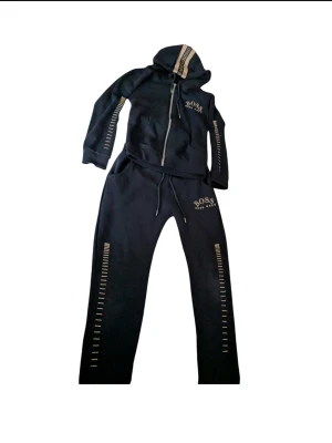 Svart set från Hugo Boss - Svart set från Hugo Boss med guldfärgade detaljer längs benen och logga på höften. Byxorna har snörning i midjan och är gjorda i mjukt bomullsmaterial. Snyggt matchande med hoodie i samma stil för en cool och avslappnad look.