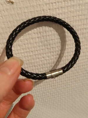 Svart flätat läderarmband - Snyggt svart armband i flätat läder med ett stilrent metallspänne. Perfekt accessoar för en enkel men cool look. Passar både till vardags och när du vill addera lite edge till outfiten.