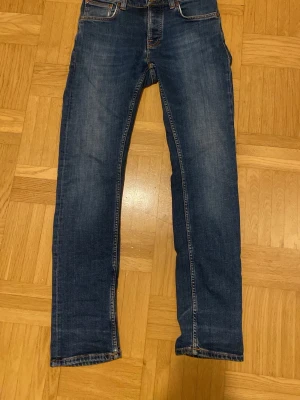 Blå jeans från Nudie Jeans Co - Säljer ett par klassiska blå jeans från Nudie Jeans Co med orangea sömmar och snygga detaljer på bakfickorna. Modellen har rak passform och normal midja, tillverkade i slitstarkt bomullsmaterial. Perfekt för dig som gillar stilrena och tidlösa jeans.