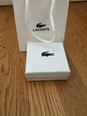 Lacoste silverfärgat armband - Säljer ett stilrent armband från Lacoste i silverfärgad metall. Armbandet har rektangulära länkar med ingraverade detaljer och den klassiska krokodilloggan. Kommer i original ask och tygpåse med Lacoste-tryck.