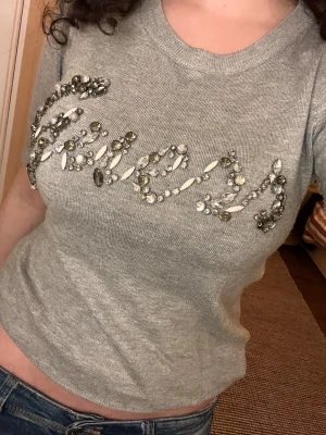 Grå stickad tröja från Guess med pärlor - Supersnygg grå stickad tröja från Guess med  pärlor och stenar som bildar loggan på bröstet. Rund halsringning och kortärmad modell. 