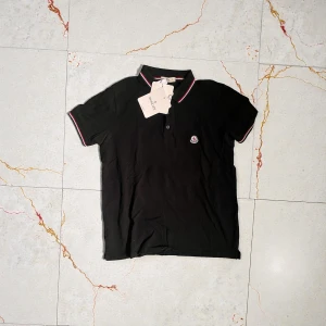 Svart Moncler piké med rödvit kant - Säljer en svart pikétröja från Moncler med klassisk krage och knappslå. Pikén har röda och vita ränder på krage och ärmslut samt Moncler-logga på bröstet. Perfekt till jeans eller shorts för en clean look.