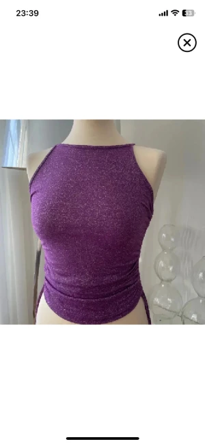 Glittrig lila linnetopp - Säljer en glittrig lila linnetopp med smala axelband och tight passform. Toppen har en snygg, rak halsringning och är tillverkad i ett stretchigt syntetmaterial som skimrar i ljuset. Perfekt för att sticka ut med en färgstark look.