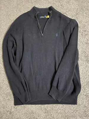 Polo Ralph Lauren half zip - Svart långärmad tröja från Polo Ralph Lauren med half zip och klassisk krage. Tröjan har ett diskret blått broderat logomärke på bröstet och är stickad i mjukt material. Perfekt för chill höst- och vinterdagar.