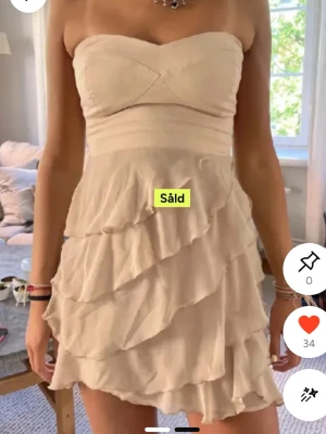 Beige axelbandslös volangklänning - Supersöt beige axelbandslös klänning med flera lager volanger och omlott-detaljer. Kort modell som ger en fräsch och somrig vibe. Perfekt för dig som gillar romantisk och feminin stil. Materialet känns lätt och luftigt.