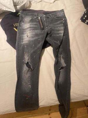 Svarta Dsquared2 jeans med färgstänk - Säljer ett par svarta jeans från Dsquared2 med coola färgstänk i vitt och rött, slitna detaljer och hål på knäna. Jeansen har klassisk femficksdesign, silverfärgad dragkedja och röda sömmar på insidan. Tillverkade i Italien och har en snygg streetstil.