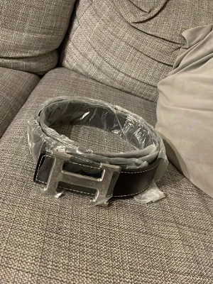 Svart bälte med H-spänne - Snyggt svart bälte med ett stort H-format metallspänne och vita kontrastsömmar längs kanterna. Bältet har en stilren och modern look som passar till jeans eller kostymbyxor. Perfekt accessoar för att lyfta din outfit.