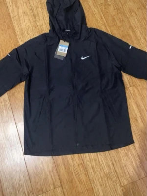 Svart vindjacka från Nike med huva - Svart vindjacka från Nike med klassisk logga på bröstet, hel dragkedja och praktisk huva. Jackan har en clean look och är tillverkad i lätt polyester som passar perfekt när det blåser. Snygg och enkel design med långärmade ärmar. Pris kan diskuteras!!
