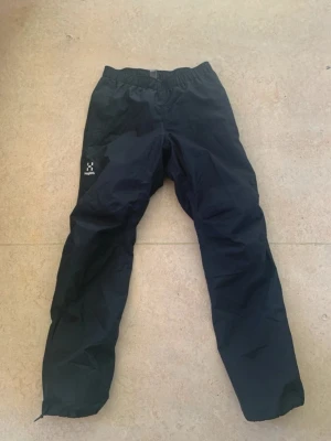 Svarta täckbyxor från Haglöfs herr S - Svarta täckbyxor från Haglöfs i herrmodell, storlek S. Byxorna har elastisk midja och är gjorda i slitstark syntet som står emot väta och vind. Perfekta för utomhusaktiviteter under kyliga dagar. Diskret Haglöfs-logga på benet.