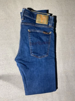 Blå jeans från Nudie Jeans - Snygga blå jeans från Nudie Jeans med klassisk femficksdesign och kontrastsömmar. Jeansen har en mörkblå tvätt och detaljerad brodering på bakfickan samt en läderpatch med logga i orange. Perfekt för dig som gillar stilrena och tidlösa jeans.